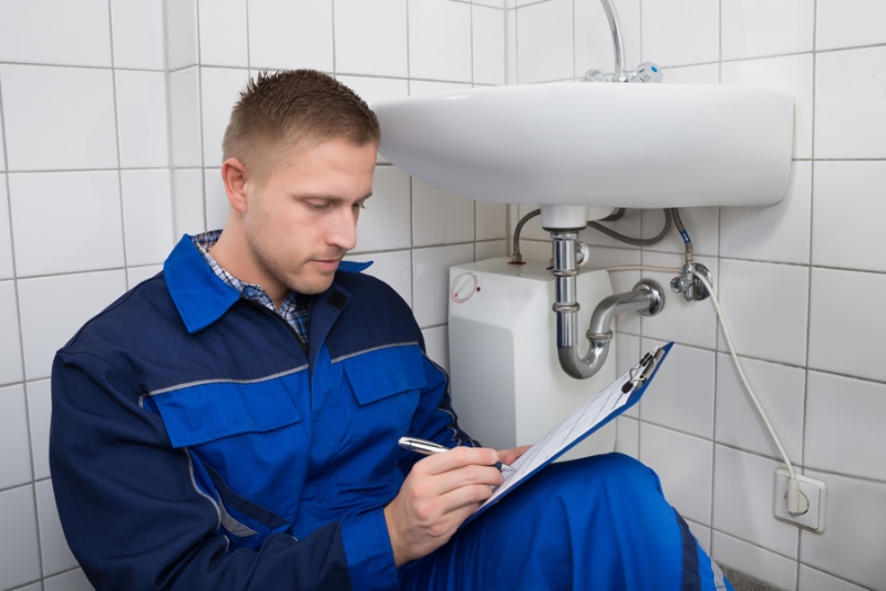 Technician writing on a clipboard sitting down near a sink. The tech is writing down findings from plumbing inspection.
